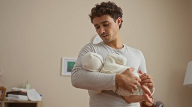 Genç İspanyol bir adam yatak odasında bebeğini kucağında tutuyor sevgi dolu aile bağlarını gösteriyor ve yanında bebek beşiği olan bir evde sıcaklığını gösteriyor.