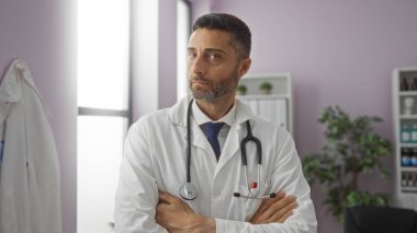 Kendine güvenen erkek doktor modern bir klinikte kollarını kavuşturarak duruyor. Doğal ışık ve tıbbi ekipmanlarla profesyonel bir sağlık ortamının altını çiziyor..