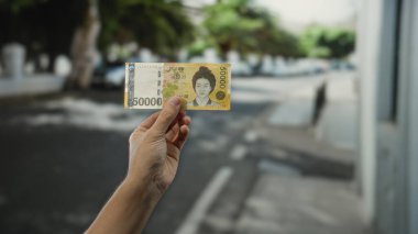 Güneşli bir günde şehir sokaklarında Güney Kore banknotunu elinde tutan adam, bulanık arka planda modern bir şehir ortamında para gösteriyor..