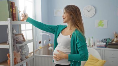 Bebek oyuncakları ve temel ihtiyaçlarla çevrili karyolasıyla bebek odasında selfie çeken hamile bir kadın evde neşeli anlar yakalıyor..
