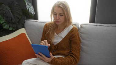 Sıcak bir apartman dairesinde kanepede oturan bir kadın tablet kullanıyor, kalın bir kazak giyiyor, yumuşak yastıklar ve bitkilerle çevrili, odaklanmış ve rahatlamış..