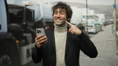 Genç adam gülümserken elinde akıllı telefonuyla şehir sokaklarında spor ceket giyerek gülümsüyor. Mutluluk, canlı kentsel yaşam tarzı, yakışıklı bir portre..