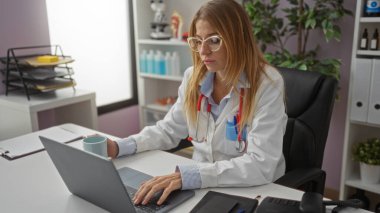 Modern klinikte laptopta çalışan doktor sarı saçlı, beyaz önlüklü, steteskoplu, gözlüklü, ciddi ifadeli, tıbbi ekipmanlarla çevrili, görevine odaklanmış..