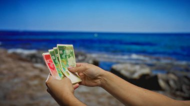 Güneşli bir günde sahil kenarında İsrail şekeli banknotları tutan adam kumsaldaki iş yatırımını ve finansal planlamayı simgeliyor..