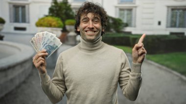 İspanyol adam elinde Polonya parası tutarken gülümsüyor ve kenar mahalle ortamında güveni ve refahı somutlaştırıyor..