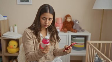 Şişeye dikkatle odaklanan bir kadın rahat bir yatak odasında elinde bir telefonla bebek dekoruyla anneliği ve teknolojiyi uyumlu bir şekilde harmanlıyor..