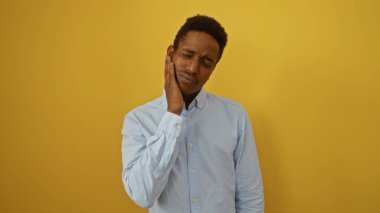 Genç adam parlak sarı bir arka plana karşı duruyor, açık mavi bir tişört giyiyor ve düşünceli bir ifade sergiliyor, modern tarzı ve kendine güveni somutlaştırıyor..