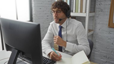 Ofiste kulaklık takan bir adam, iyi aydınlatılmış bir masada notlar alırken, profesyonel ve düşünceli bir çalışma ortamını iletirken çevrimiçi bir toplantıya katılıyor..