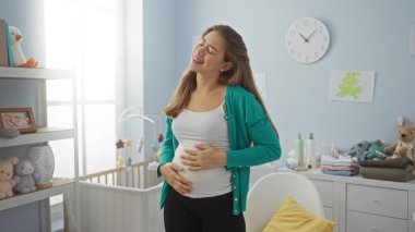 Hamile kadın, bebek eşyaları ve bebek odası dekoruyla çevrili, rahat bir yatak odasında dururken nazikçe gülümsüyor. Huzurlu bir evde yaklaşan anneliğin mutluluğunu kucaklıyor..