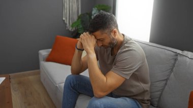 Genç adam rahat bir oturma odasında kanepede otururken düşünceli görünüyor, elleri düşünceye sıkışmış, günlük giysiler giyiyor, evdeki iç gözlem duygusunu vurguluyor..