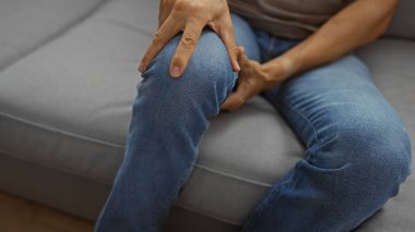 Oturma odasındaki kanepede oturan genç adam dizini tutuyor, kot pantolon giyiyor, rahat bir apartman dairesinde, rahatsızlık ya da acıyı işaret ediyor..