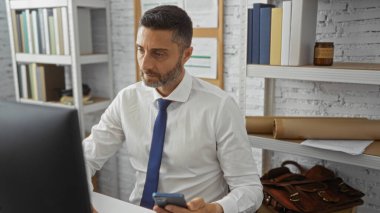 Parlak ofiste çalışan bir adam telefonu bilgisayar ekranına odaklıyor. Etrafı kitaplarla çevrili. Kapalı alandaki profesyonel ayarlar..