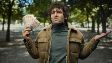 Ağaçlarla çevrili açık şehir ortamında sorgulayıcı bir ifadeyle elinde Türk lirası tutan bir adam İspanyol özellikleri sergiliyor ve sıcak bir ceket giyiyor..