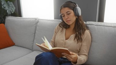 Kadın kitap okurken rahat bir oturma odasında kulaklıkla müzik dinliyor, rahat bir iç mekan sergiliyor..