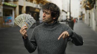 Hispanik bir adam şehir sokaklarında Amerikan dolarlarını sergiliyor, özgüvenle hareket ediyor ve açık bir şehir ortamında banknotların değerini vurguluyor..