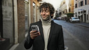 Kıvırcık saçlı ve sakallı genç adam şehir caddesinde akıllı telefona bakıp neşeli yüz ifadesini sergilerken gülümsüyor..