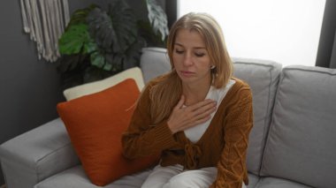 Kapalı bir evde, rahat bir oturma odasında oturan, kahverengi hırkalı, turuncu yastıklı ve arka planda yeşil bitkilerle rahatsız olan bir kadın..