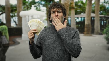 Norveçli kronu elinde tutan adam palmiye ağaçlarıyla çevrili düşünceli bir ifadeye sahip bir şehirde açık havada para yatırıyor..