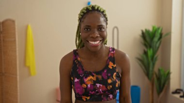 Spor malzemeleriyle çevrili, modern bir spor salonunda, saç stili örülmüş, canlı spor kıyafetleri içinde, parlak bir şekilde gülümseyen bir kadın..