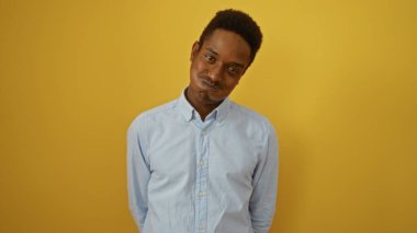 İzole edilmiş sarı arka planda, yakışıklı ve çekici özelliklerini vurgulayan bir stüdyo ortamında resmedilmiş mavi bir gömlek giyen genç adam neşeli bir ifade sergiliyor.