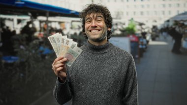 Czech Koruna banknotlarını elinde tutarken gülümseyen İspanyol adam hareketli bir şehir terasında, restoranları ve kafeleri olan.
