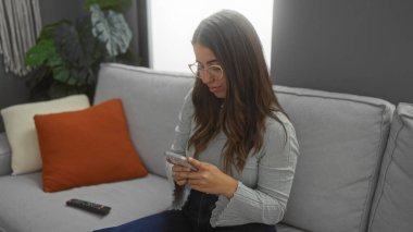 Oturma odasında oturan kadın rahat bir yaşam tarzına odaklanmış akıllı telefon kullanıyor rahat dekorlu ve yumuşak ışıklandırmalı modern bir dairede ılık bir atmosfer yaratıyor..