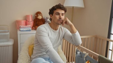 Genç İspanyol bir adam, yumuşak oyuncaklar ve sıcak aydınlatmalarla çevrili, rahat bir yatak odasında, bebek beşiğinin yanında, düşünceli bir şekilde oturuyor..