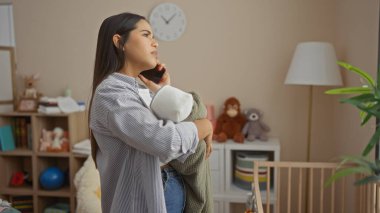 Yatak odasında oyuncaklarla ve beşiğiyle telefonda konuşurken kucağında bebek tutan bir kadın, evde ilgili ebeveynlik atmosferi sergiliyor..