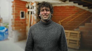 İnşaat alanındaki genç adam kendine güveni tam, gri bir kazak giyiyor, tuğla duvarlar ve ahşap paletler ile çevrili..