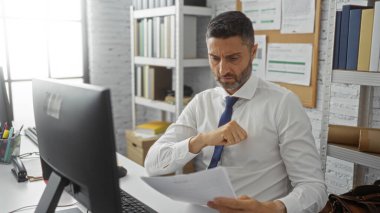 Ofisteki adam belgeleri ciddi şekilde analiz ediyor. Etrafı raflarla çevrili. İç kısımları düzenli bir bilgisayar ve beyaz gömlekli, mavi kravatlı..