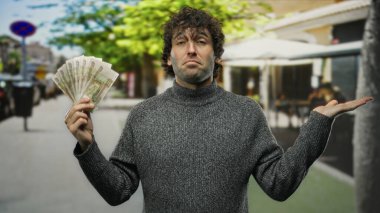 Gri kazaklı bir adam, şehir arka planı bulanık ve canlı yeşil ağaçlarla çevrili, düşünceli bir ifadeyle bir şehir caddesinde Rus rublesi tutuyor..