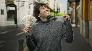 Hispanik bir adam şehrin dışında elinde Rus rublesi tutarken gülümseyip yukarıya doğru işaret ederek finansal başarı ya da zenginlik öneriyor..