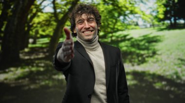 Güneşli bir parkta ışıl ışıl gülümseyen yakışıklı İspanyol genç adam, güneşli bir günde açık hava ortamını çevreleyen yemyeşil ağaçlarla misafirperver bir tokalaşma uzattı.