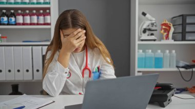 Kadın doktor klinikte elleri yüzü koyun, steteskop takmış, tıbbi ekipmanlarla çevrili, profesyonel sağlık hizmetlerinde yorgunluk belirtileri gösteriyor..