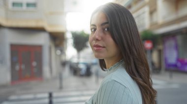 Uzun esmer saçlı bir kadın, şehir sokağında ayakta duruyor. Hoş bir gülümseme sergiliyor. Gün boyunca etrafındaki hayat ve atmosferi yansıtıyor..