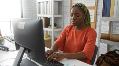 Modern ofis ortamında bilgisayar başında çalışan, turuncu giyinen, odaklanmış ifade sergileyen, saçlarını ören bir kadın..
