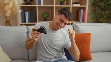 Genç adam rahat bir oturma odasında video oyunları oynuyor, elinde bir kumanda, rahat bir koltukta oturuyor ve etrafında kitap rafları ve bitkiler var..