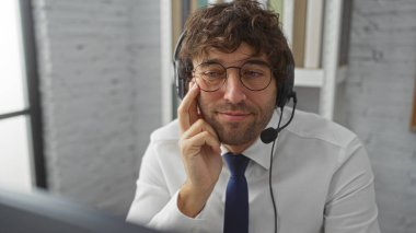 Gözlüklü ve kulaklık takan genç bir adam bir ofis odasında oturuyor, odaklanmış ve profesyonel görünüyor, modern bir çalışma ortamını örnek alıyor, gündelik ama özenli bir tavırla..