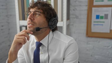 Kulaklıklı genç adam, belgelerle ve teknolojiyle çevrili modern bir ofis ortamında düşünceli bir şekilde, verimli bir çalışma ortamı öneriyor..