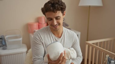 Sıcak bir yatak odasında bebeğini tutarken gülümseyen genç bir adam, şefkatli bir baba ve mutluluk anı sergiliyor..