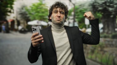 Parkta gururla duran, elinde akıllı bir telefon tutan balıkçı yaka ve ceket giyen, özgüven gösteren ve arka planda bulanık yapraklarla etkileyici bir jest yapan bir adam.