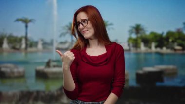 Parkta gözlüklü ve kızıl saçlı bir kadın, kırmızı bir süveter giyiyor, neşeli görünüyor ve başparmağıyla bir çeşme ve yeşillik zeminine işaret ediyor..