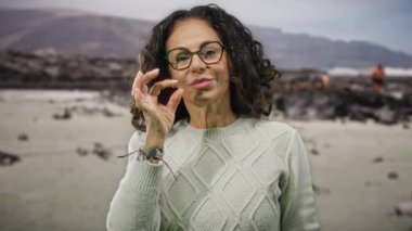 Kıvırcık saçlı bir kadın, deniz kenarında bir sahilde, gözlük ve süveter giyerken jest yapıyor. Manzaralı bir arka planı olduğunu söylüyor..