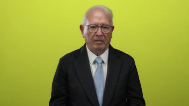 Takım elbiseli kıdemli bir adam, elleri izole edilmiş sarı arka plan duvarının üzerinde, düşünceli bir ifadeyi canlandırıyor..