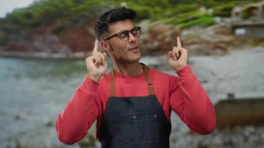 Gözlüklü ve önlüklü genç adam sahilde neşeyle gülümsüyor manzaralı bir arka planda.