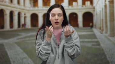 Esmer saçlı bir kadın, üniversite kampüsünde sürpriz yapıyor. Kemerleri açık havada eğitim ortamında günlük gri kapüşonlu giyiniyor..