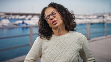 Kıvırcık saçlı İspanyol kadın arka planda teknelerle deniz kenarında rahatsızlık gösteriyor. Açık havada süveter giymesi mide ağrısı ya da rahatsızlık hissi veriyor..