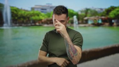 Dövmeli genç adam, güneşli bir park atmosferinde bir fıskiyenin ve yemyeşil bir yeşilliğin yer aldığı tetikte ve uyarıda bulunuyor..