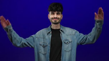 Kot ceketli genç adam canlı mavi arka planda gülümsüyor, kollarını kaldırarak pozitif ve kendinden emin bir ifade sergiliyor, yakışıklı ve çekici özellikleri vurguluyor..