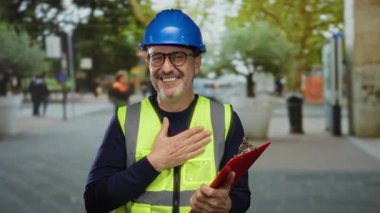 Şehir sokaklarında gülümseyen bir adam, arka plandaki meslektaşlarıyla pano tutarken kasket ve güvenlik yeleği giyerek neşeli profesyonel sahneleri gösteriyor..
